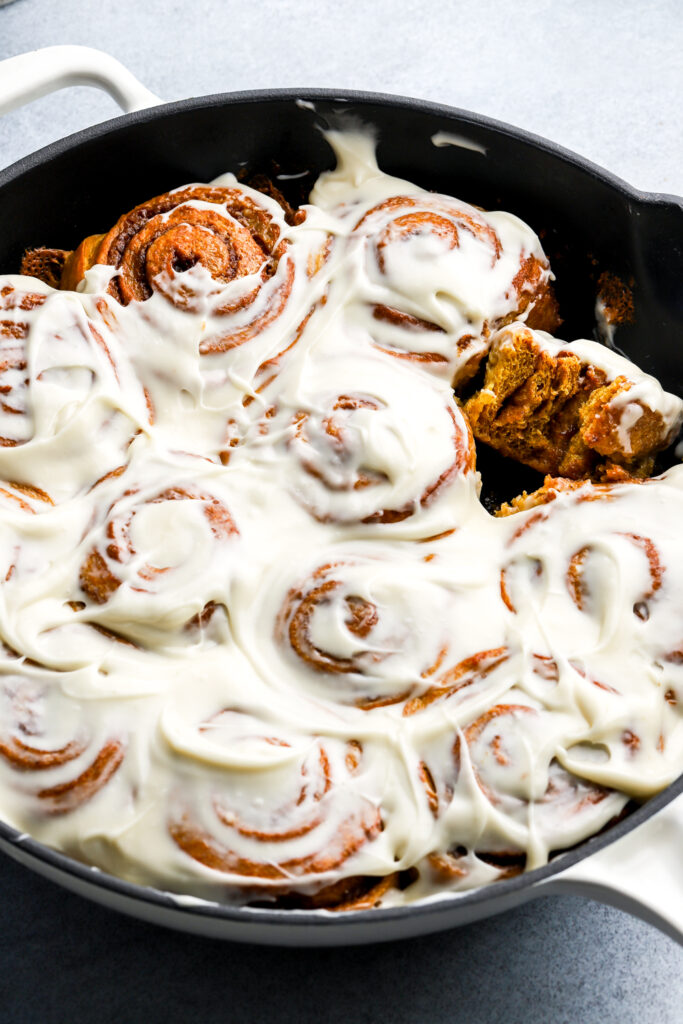 12 pumpkin cinnamon rolls covered in icing still in the pan with one roll tilted and bitten